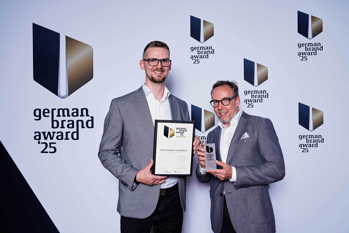 2 Männer lächeln in die Kamera und halten die Urkunde und Aufsteller des German Brand Awards 2025 für HörPartner in den Händen